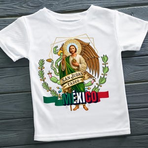 May include: White t-shirt featuring a graphic of San Judas Tadeo with a halo, wings, and a staff. The design includes the words "SAN JUDAS TADEO" and "MEXICO" with the Mexican flag colors.