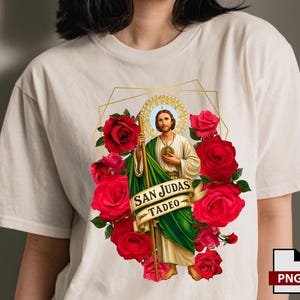May include: Cream-colored t-shirt featuring a religious graphic of San Judas Tadeo surrounded by red roses. The image includes a banner with the text "SAN JUDAS TADEO". The design is framed by a gold geometric shape.