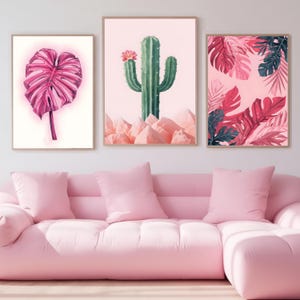 Puede incluir: Tres impresiones artísticas botánicas enmarcadas. Las impresiones presentan una hoja rosa, un cactus verde con una flor rosa y hojas rosas y verde oscuro. Las impresiones se muestran sobre un sofá rosa con cojines rosas.
