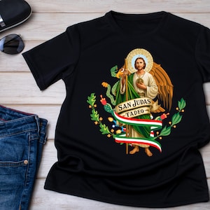 May include: Black t-shirt featuring a religious image of San Judas Tadeo with a halo, wings, and a staff. The design includes a banner with the text "SAN JUDAS TADEO" and a Mexican flag-inspired ribbon. The shirt is displayed on a wooden surface.