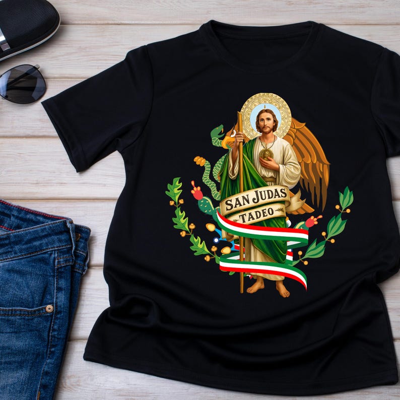 May include: Black t-shirt featuring a religious image of San Judas Tadeo with a halo, wings, and a staff. The design includes a banner with the text "SAN JUDAS TADEO" and a Mexican flag-inspired ribbon. The shirt is displayed on a wooden surface.