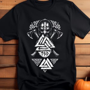 Puede incluir: Camiseta negra con un gráfico vikingo blanco. El diseño incluye un casco, hachas y un símbolo triangular con las letras "TSD". La camiseta está hecha de un material suave.