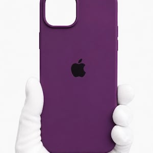 Pode incluir: Uma capa de telefone roxa lisa com um logotipo da Apple preto. A capa tem recortes para a câmera e botões. A capa do telefone é segurada por uma mão enluvada.
