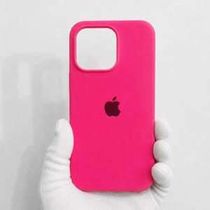 Pode incluir: Uma capa de telefone rosa brilhante com um logotipo escuro. A capa tem um recorte para a câmera e é segurada por uma mão enluvada. O fundo é branco liso.