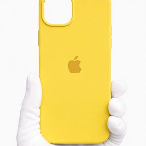 Pode incluir: Uma capa de telefone amarelo brilhante com um recorte para a câmera e um logotipo da Apple dourado. A capa é segurada por uma mão enluvada em um fundo branco.