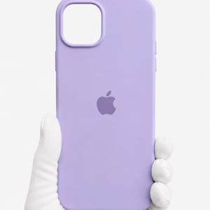 Pode incluir: Uma capa de telefone de cor lavanda com um recorte para a câmera e o logotipo da Apple. A capa é feita de um material liso e foi projetada para proteger o telefone. A capa é segurada por uma mão enluvada.