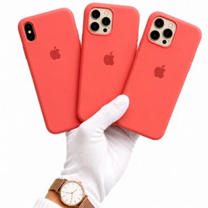 Pode incluir: Três capas de telefone cor-de-rosa com o logotipo da Apple são seguradas em uma mão enluvada. Um relógio dourado com uma pulseira marrom é visível no pulso. As capas são para diferentes modelos de iPhone.