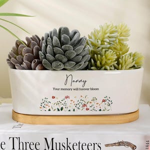 Op de afbeelding: Een witte rechthoekige keramische plantenbak met een houten voet, gevuld met drie kunstmatige vetplanten. De plantenbak bevat de tekst Nanny, Your memory will forever bloom en een bloemenpatroon. Het staat op een boek getiteld The Three Musketeers.