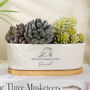 May include: A white rectangular ceramic planter with a wooden base, featuring a black line-art cat illustration, the text "NEVER FORGOTTEN" and the name "Daniel" printed on the front, containing two artificial succulent plants.