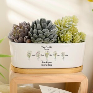 May include: A white rectangular ceramic planter with a wooden base, containing three succulents. The planter features the text "Mrs Smith 2024-2029" above a row of five small floral illustrations with the names Amelia, Emily, Olivia, Tracy, and Sophie.