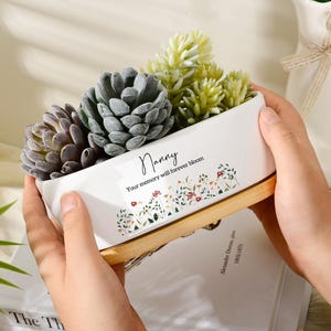 Op de afbeelding: Een rechthoekige witte keramische plantenbak met een bamboe voet, gevuld met diverse kunstmatige vetplanten. De plantenbak is voorzien van de tekst Nanny, Your memory will forever bloom en een kleurrijke bloemenprint op de voorzijde.