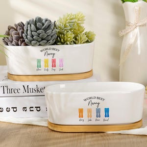 May include: Two white oval ceramic planters with bamboo bases. Each planter features the text "WORLD BEST Nanny" above four colorful illustrations of rain boots, each labeled with a name. One planter holds green and grey succulents.