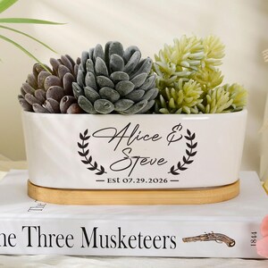 May include: A white oval ceramic planter with a wooden base, containing three succulent plants. The planter features black text reading Alice & Steve and Est 07.29.2026, framed by two laurel branches. It sits atop a book titled The Three Musketeers.