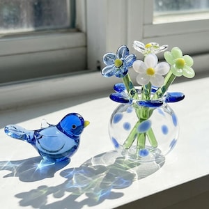 May include: A small blue glass bird figurine positioned next to a clear glass vase with blue polka dots containing three glass flowers in blue, white, and light green, each featuring a small yellow center on thin green glass stems.