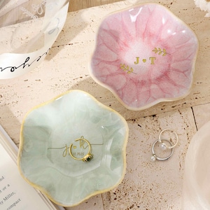 May include: Two flower-shaped ceramic trinket dishes, one in soft pink and one in pale green, each featuring gold-colored lettering and leaf accents. The pink dish displays the initials J and T, while the green dish holds a gold-toned ring.