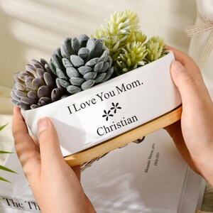Puede incluir: Una maceta rectangular de cerámica blanca con base de madera clara, que contiene varias plantas suculentas artificiales. El frente de la maceta presenta el texto impreso en negro "I Love You Mom." seguido de dos pequeños iconos de flores y el nombre "Christian."
