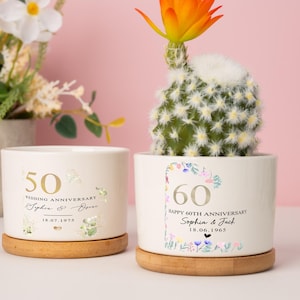Può includere: Due vasi in ceramica bianchi con basi in legno, ciascuno con un cactus. Un vaso mostra "50 WEDDING ANNIVERSARY" con una data, l'altro recita "60 HAPPY 60TH ANNIVERSARY" con una data. Il cactus nel secondo vaso ha un fiore arancione.