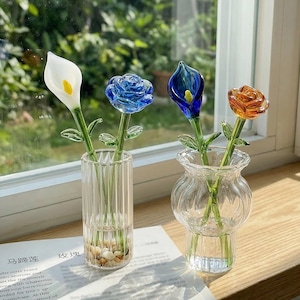 May include: Glass flower sculptures in a vase. The flowers include a white calla lily, a blue rose, a blue calla lily, and an orange rose. The flowers are in two clear glass vases. The vases are on a wooden surface near a window.