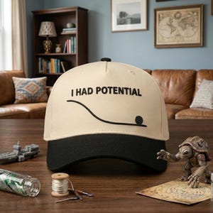 Pode incluir: Boné de beisebol bege com aba preta. O boné apresenta o texto "I HAD POTENTIAL" acima de uma linha preta e um ponto preto. O chapéu é exibido em uma superfície de madeira, com uma figura de tartaruga em miniatura e outros objetos.