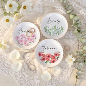 May include: Three white ceramic ring dishes with gold rims, each with a floral design and a name: Carolyn, Beverly, and Fabiana. The Carolyn dish has pink flowers, Beverly has snowdrops, and Fabiana has red poppies. Gold rings and a silver necklace are also present.