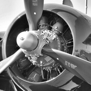 May include: A black and white close-up photo of an airplane propeller and engine. The propeller is made of metal and has a black and white paint scheme. The engine is visible behind the propeller and has a complex design with many parts.