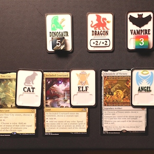 May include: A collection of Magic: The Gathering cards and creature tokens. Tokens include a dinosaur, dragon, and vampire. Cards feature an elf, angel, and cat. The tokens have white borders and are on a dark surface.