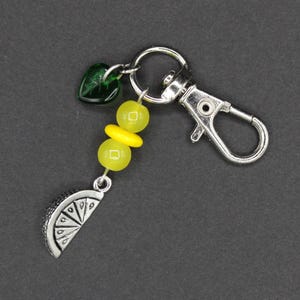 May include: A silver-toned keychain with a lobster clasp, featuring a charm with a lemon slice design, yellow beads, and a green heart-shaped bead. The keychain is on a dark gray background.