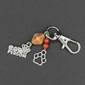 May include: A silver keychain with a lobster clasp, featuring charms and beads. Charms include a paw print and the words "CAT PERSON". The beads are brown and red. The keychain is designed for cat lovers.