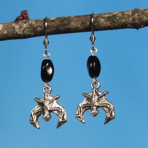 May include: Silver bat earrings with black beads hanging from a branch against a blue sky background.