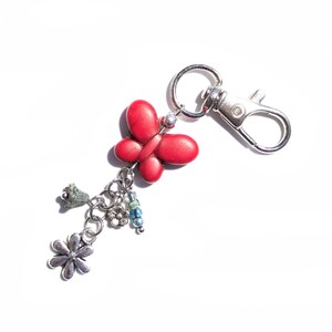 May include: A red butterfly charm keyring with a silver clasp. The keyring features a red butterfly, a small flower, a green charm, and blue beads. The keyring is on a white background.