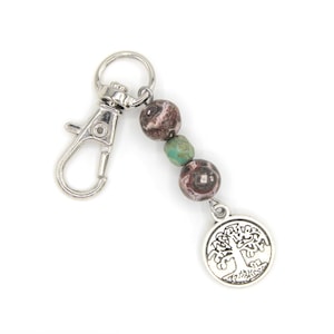May include: A silver-toned keychain with a lobster clasp, featuring three beads in shades of brown and green. A round charm with a tree design hangs below the beads. The keychain is on a white background.