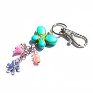 May include: A turquoise butterfly charm keychain with a silver clasp. The butterfly is accented with gold and has a cluster of pink and silver charms, including a flower and beads. The keychain is on a white background.
