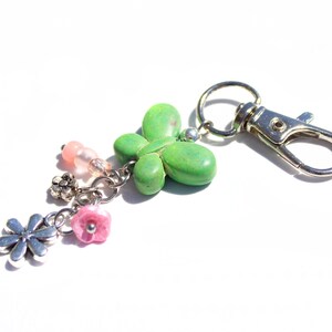 May include: A decorative keyring with a silver clasp and ring. The keyring features a green, four-leaf clover charm, along with pink and silver flower charms and pink beads. The charms are attached with silver chains.