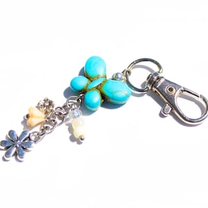 May include: A silver-coloured keyring featuring a turquoise stone flower design and dangling charms. The charms include small flowers and beads. The keyring has a lobster clasp for easy attachment.