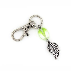 May include: A silver-colored keychain with a lobster clasp, a green and clear bead, and a decorative leaf charm. The leaf charm has intricate detailing. The keychain is on a white background.