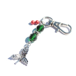 May include: A silver-coloured keyring with a lobster clasp, featuring a holly leaf charm and green faceted beads. The keyring also includes red beads and is set against a white background. A festive accessory.