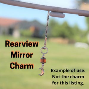 May include: A rearview mirror charm with a silver chain, a lobster clasp, and decorative beads. The charm features a small, crescent moon pendant and the words "Rearview Mirror Charm". The background is blurred, with green foliage and a building.