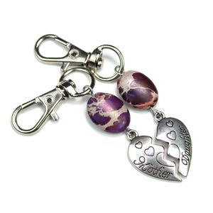 May include: A keychain featuring two silver clasps, speckled purple and brown beads, and a split heart charm. One half of the heart says "Mother," the other says "Daughter." The item is set against a white backdrop.