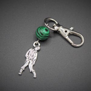 May include: A silver-colored keychain with a zombie charm and a green and black malachite bead. The keychain has a lobster clasp for easy attachment to bags or keys. The zombie charm is detailed and stylized.