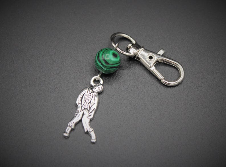 May include: A silver-colored keychain with a zombie charm and a green and black malachite bead. The keychain has a lobster clasp for easy attachment to bags or keys. The zombie charm is detailed and stylized.