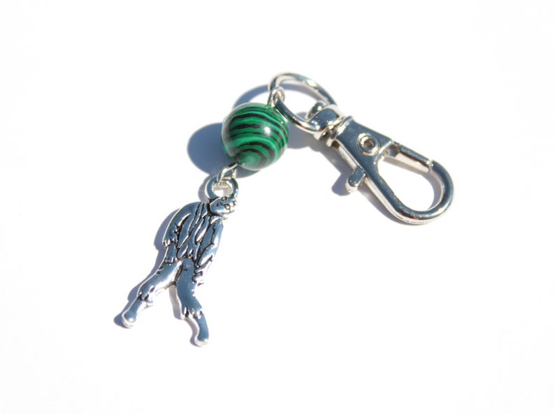 May include: A silver-toned keychain with a malachite green and black bead and a charm shaped like a mythical creature. The keychain has a silver clasp for easy attachment.