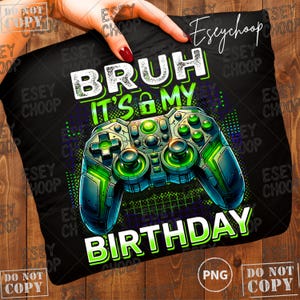 Peut inclure: Coussin noir avec un motif de manette de jeu vidéo vert néon et bleu. Le texte "BRUH IT'S MY BIRTHDAY" est en blanc et vert. Le coussin est tenu par une main avec du vernis à ongles rouge.