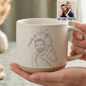 May include: Cream-colored ceramic mug with a sketch of a man and child, with the text "Daddy & Chloe." The mug has a light brown base and a large handle. A small photo with the text "UPLOAD PHOTO" is in the upper right corner.