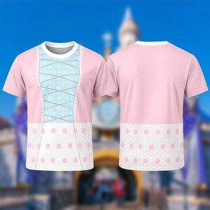 Puede incluir: Camiseta rosa con un dise&ntilde;o que simula un vestido. La parte delantera tiene un detalle de cors&eacute; azul claro con ribete de encaje blanco y un estampado de lunares blancos. La parte trasera es rosa liso con un estampado de lunares blancos en la parte inferior.