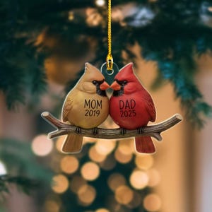 Puede incluir: Adorno navide&ntilde;o decorativo con dos aves cardenales posadas en una rama. Una es beige, la otra roja. La beige tiene la inscripci&oacute;n "MOM 2019", la roja "DAD 2025". Colgando de un hilo dorado, sobre un fondo borroso.