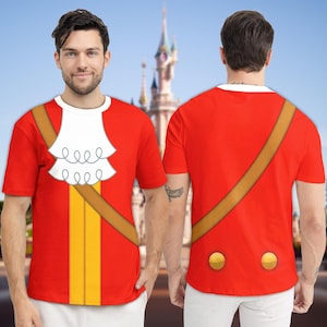May include: Red t-shirt with a white ruffled collar, brown strap, and yellow stripe design. The back of the shirt features a brown strap and two gold buttons. The shirt is a costume-themed design.