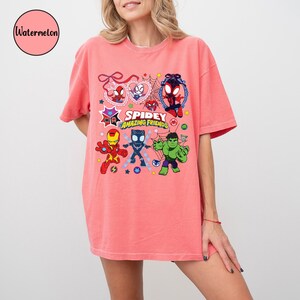 K&ouml;nnte beinhalten: Korallfarbenes T-Shirt mit einer Cartoon-Grafik von Superhelden, darunter Spider-Man, Iron Man, Black Panther und Hulk. Der Text "Spidey Amazing Friends" ist ebenfalls abgebildet. Das Shirt hat kurze &Auml;rmel und eine lockere Passform.