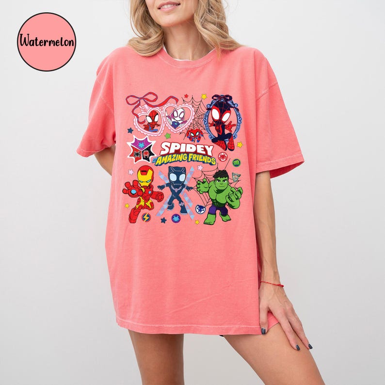 K&ouml;nnte beinhalten: Korallfarbenes T-Shirt mit einer Cartoon-Grafik von Superhelden, darunter Spider-Man, Iron Man, Black Panther und Hulk. Der Text "Spidey Amazing Friends" ist ebenfalls abgebildet. Das Shirt hat kurze &Auml;rmel und eine lockere Passform.