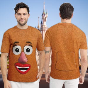 May include: A short-sleeved, orange t-shirt featuring a cartoon face with large eyes, a pink nose, and a red smiling mouth. The back of the shirt has a pocket design. The shirt is worn by a person.