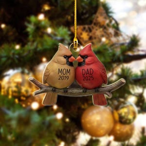 Puede incluir: Adorno navide&ntilde;o con dos cardenales posados en una rama. Un p&aacute;jaro es de color beige con la palabra "MOM" y el a&ntilde;o "2019", el otro es rojo con la palabra "DAD" y el a&ntilde;o "2025". El adorno cuelga de un hilo dorado.